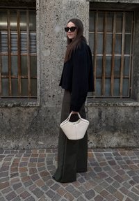 Femme en veste noire et pantalon large tenant un sac à main blanc, debout dans une rue pavée à côté d'un mur en béton avec des fenêtres grillagées.