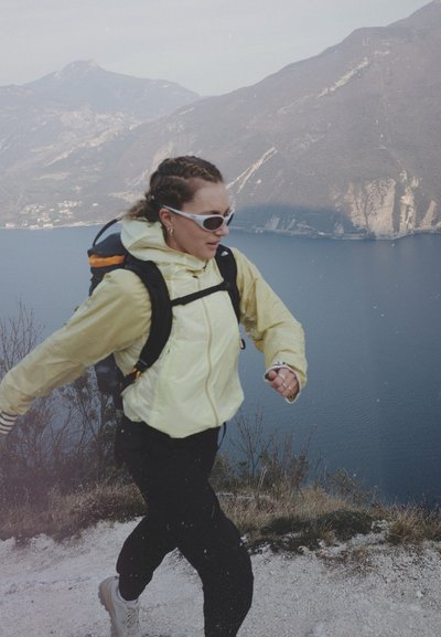 Mujer con chaqueta amarilla y gafas de sol haciendo senderismo por un sendero de montaña con vistas a un lago y acantilados escarpados.
