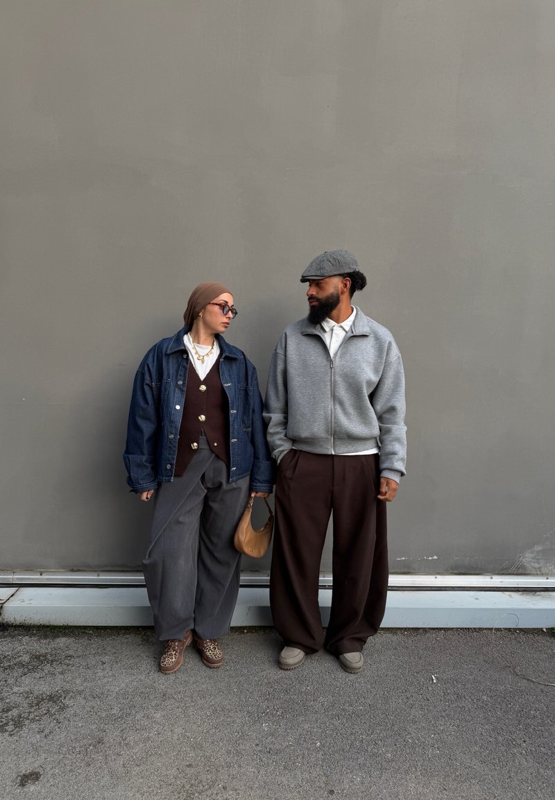 Deux personnes se tiennent contre un mur gris, portant des vêtements décontractés et amples ; l'une porte une veste en jean et un foulard sur la tête, l'autre un sweat à capuche gris zippé et une casquette.