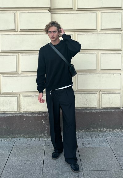 Suéter negro y pantalones sueltos negros, combinados con zapatos negros. Se lleva un bolso cruzado, con una pared texturizada de fondo.