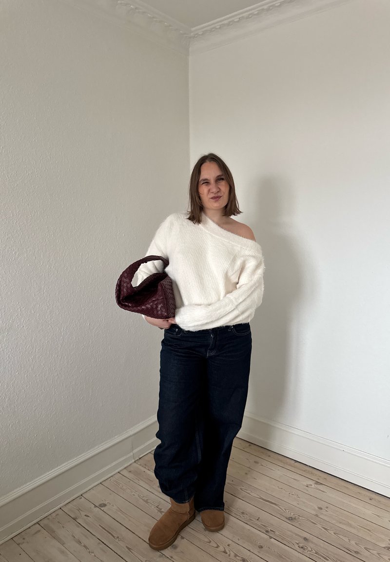 Donna che indossa un maglione bianco con le spalle scoperte, jeans scuri e stivali marroni, che tiene una grande borsa intrecciata di colore rosso scuro in una stanza spoglia.