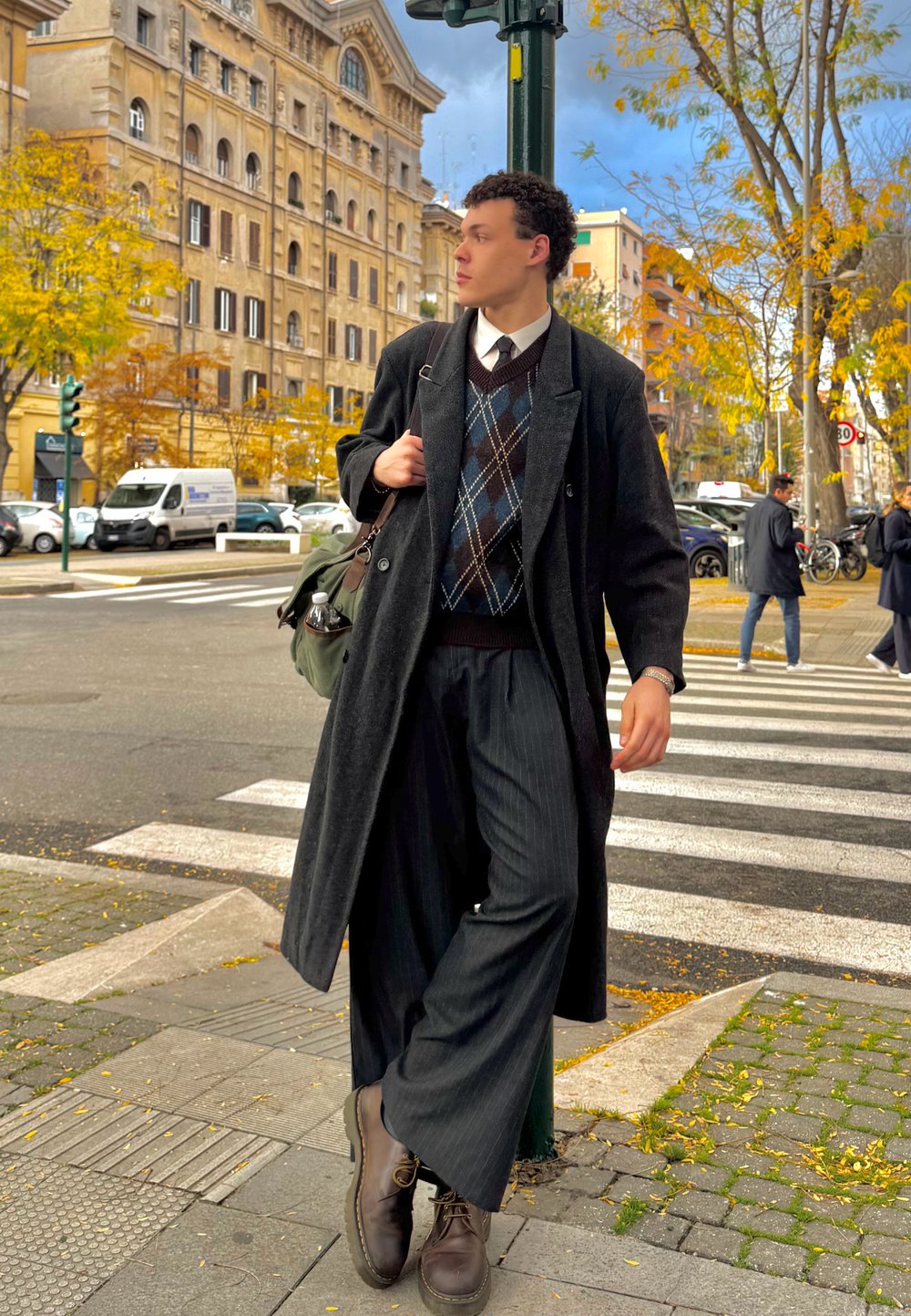 Garš melns mētelis, rakstaina brūna un zila džemperis, platai svītrainas bikses un brūni zābaki; stāv pie pilsētas pārejas ar kokiem netālu.