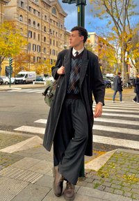 Garš melns mētelis, rakstaina brūna un zila džemperis, platai svītrainas bikses un brūni zābaki; stāv pie pilsētas pārejas ar kokiem netālu.