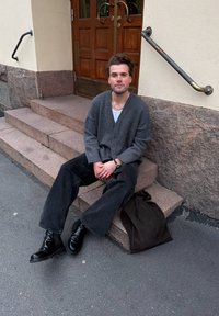 Grey cardigan over a white shirt, paired with loose black jeans and black boots. Sitting on stone steps by wooden double doors. Brown bag beside.