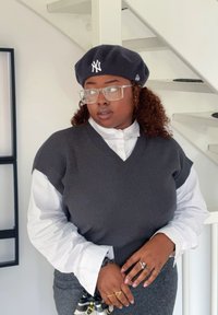 Szénszínű kötött mellény fehér gombos ing fölött, sötét barettel, amelyen logó látható, valamint gyűrűkkel és texturált anyaggal kiegészítve.