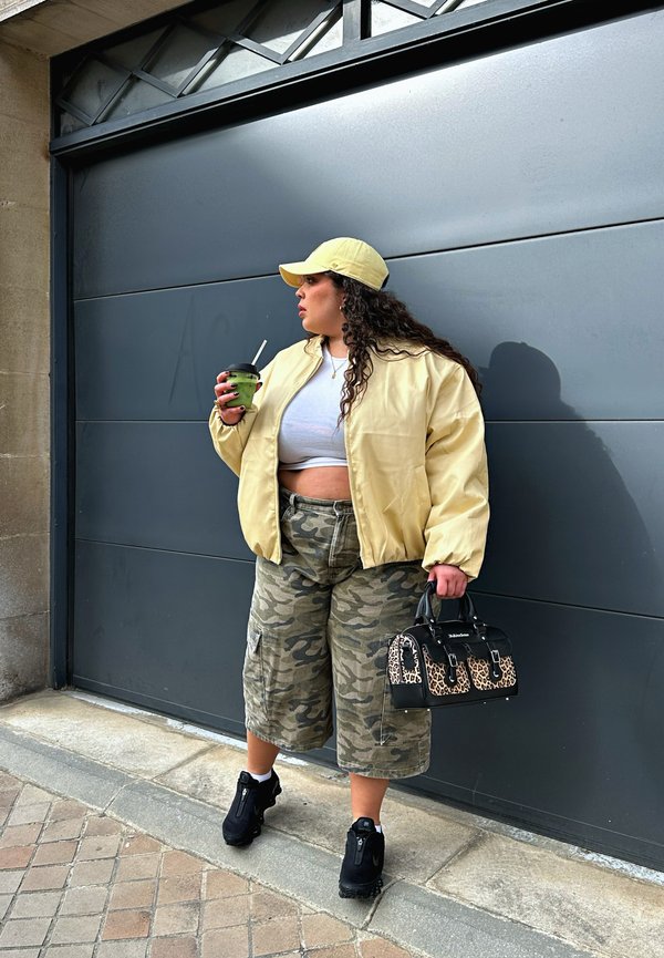 Kvinde iført gul jakke og kasket, camouflage shorts, holder en leopardprint håndtaske og en grøn kop med sugerør, står ved en grå port.