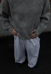 Suéter de punto a rayas grises y negras con mangas anchas, combinado con pantalones deportivos grises de ajuste holgado. Manos adornadas con anillos plateados, de pie sobre asfalto.