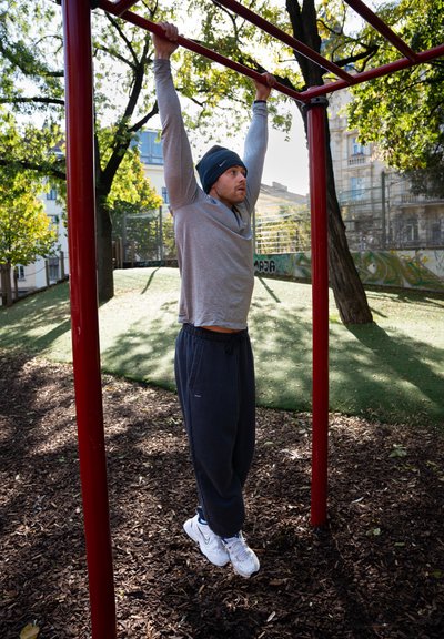 Hombre colgado de una barra de dominadas roja al aire libre, vistiendo una camiseta de manga larga gris y pantalones de chándal oscuros, con zapatillas deportivas blancas.