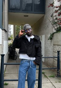 Casaco preto cropped com bordas desfiadas sobre uma t-shirt branca, combinado com jeans azul claro. A pessoa usa óculos transparentes e um gorro estampado.