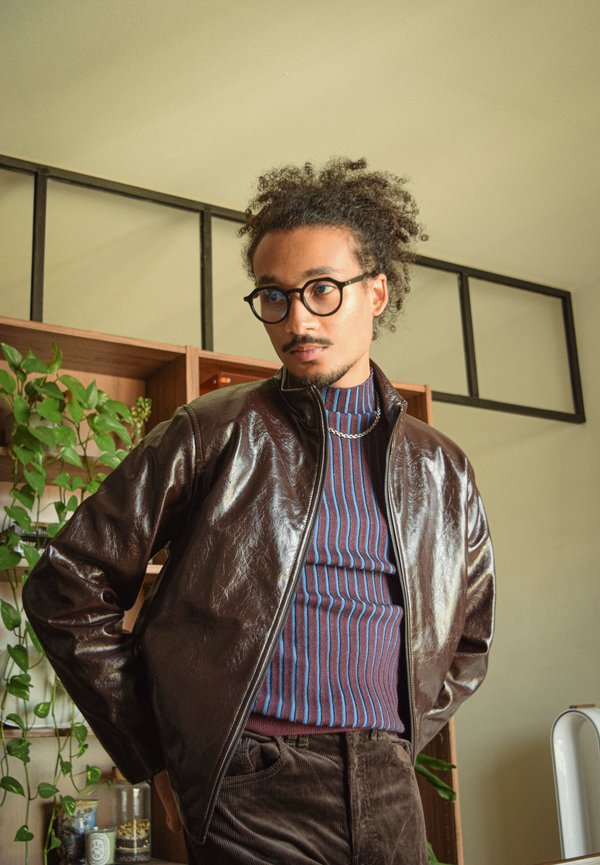 Veste en cuir marron avec une fermeture éclair, associée à un col roulé rayé. Le modèle a les cheveux bouclés et porte des lunettes, installé devant des étagères en bois.