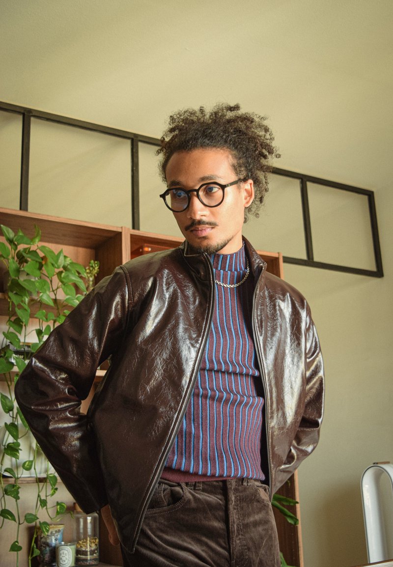 Veste en cuir marron avec une fermeture éclair, associée à un col roulé rayé. Le modèle a les cheveux bouclés et porte des lunettes, installé devant des étagères en bois.