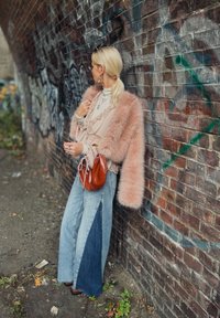 Cappotto in pelliccia finta rosa, top beige stratificato, jeans larghi in denim con pannelli laterali blu scuro e borsa in pelle marrone contro un muro di mattoni.