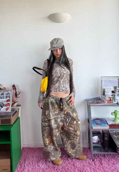 Mujer joven con pantalones de camuflaje, top marrón con estampado, gorra beige de Adidas y bolso amarillo brillante de pie en una habitación con estanterías y revistas.