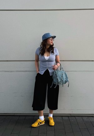 Mujer con culottes negros, top azul a cuadros, gorra azul, zapatillas amarillas y gafas, de pie contra una pared blanca sosteniendo un bolso azul claro.