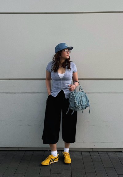 Mujer con culottes negros, top azul a cuadros, gorra azul, zapatillas amarillas y gafas, de pie contra una pared blanca sosteniendo un bolso azul claro.