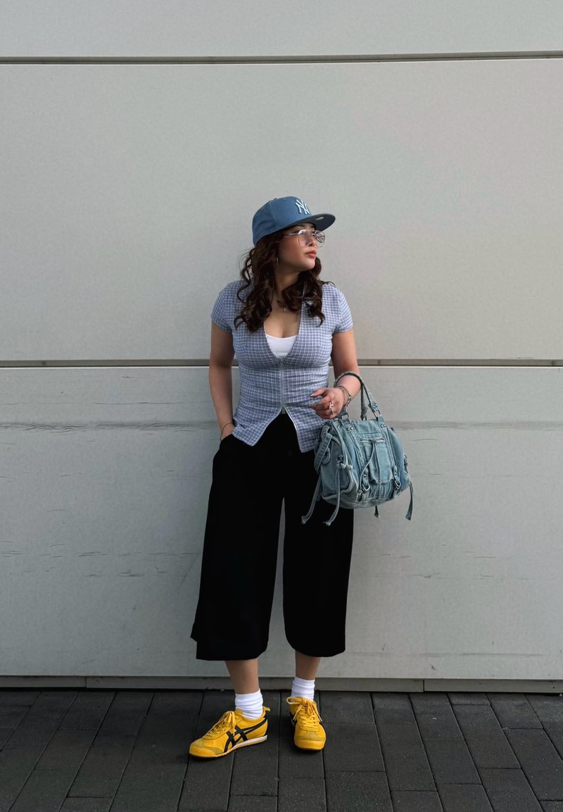 Femme en culottes noires, haut à carreaux bleu, casquette bleue, baskets jaunes et lunettes, debout contre un mur blanc, tenant un sac à main bleu clair.