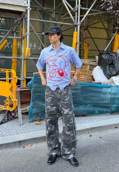 Camisa de manga corta a rayas con diseño gráfico rojo, combinada con pantalones cargo camuflados grises. Hombre de pie en una calle, rodeado de materiales de construcción.