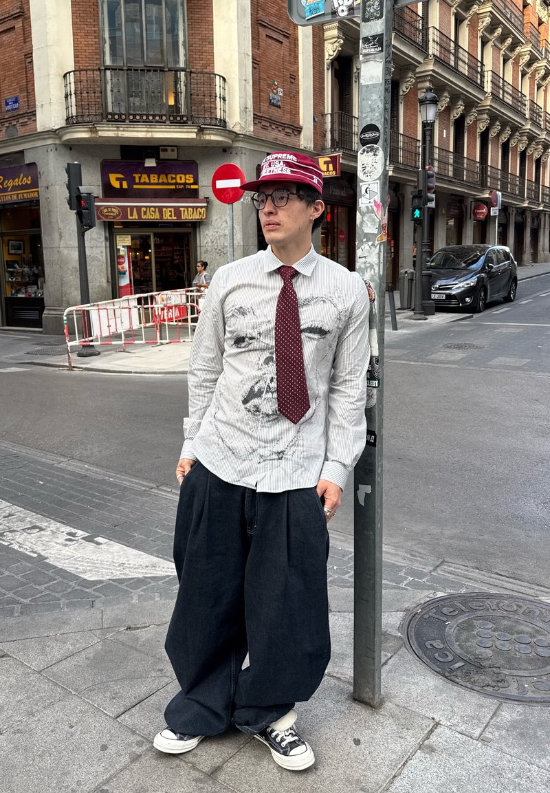 A person wearing a white striped shirt with a graphic design, a maroon tie, and oversized dark jeans, stands by a lamp post in an urban setting.