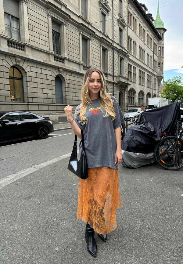 Graues übergroßes T-Shirt mit "Give a Day" Grafik, orangener Spitzenrock und schwarzen knielangen Stiefeln. Eine schwarze Umhängetasche setzt einen Kontrast.