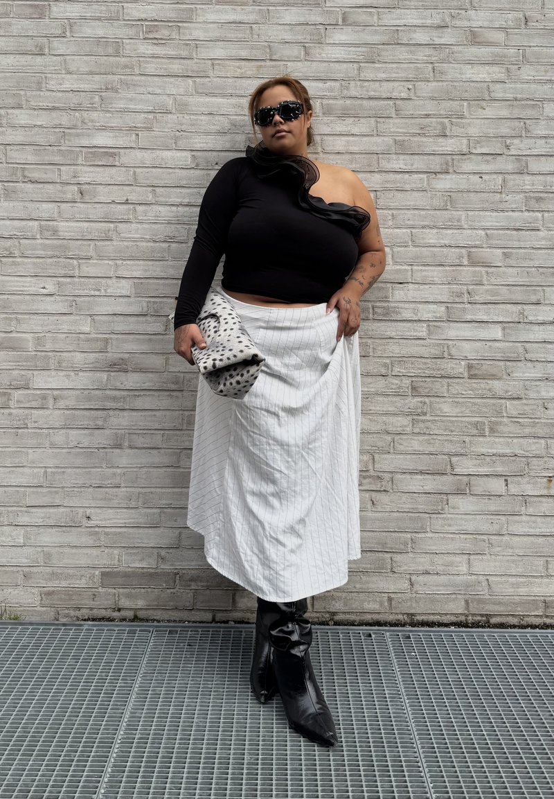 Woman wearing one-shoulder black top, white pinstriped skirt, black boots, and sunglasses, holding a spotted clutch, standing against a gray brick wall.