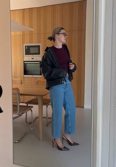 Mujer con gafas, chaqueta negra, camiseta burdeos, vaqueros azules y tacones altos burdeos, de pie en una cocina moderna con armarios y mesa de madera.