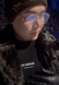 Personne portant des lunettes, un bonnet marron, un manteau en fausse fourrure noire et une chemise noire avec le texte blanc « HAN KJØBENHAVN SEARCHING FOR SHADOWS » dans une lumière tamisée.