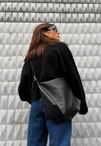 Czarna skórzana torba na ramię o luźnym kroju i minimalnych dodatkach. Modelka nosi czarny sweter i niebieskie dżinsy na tle teksturowanej szarej ściany.