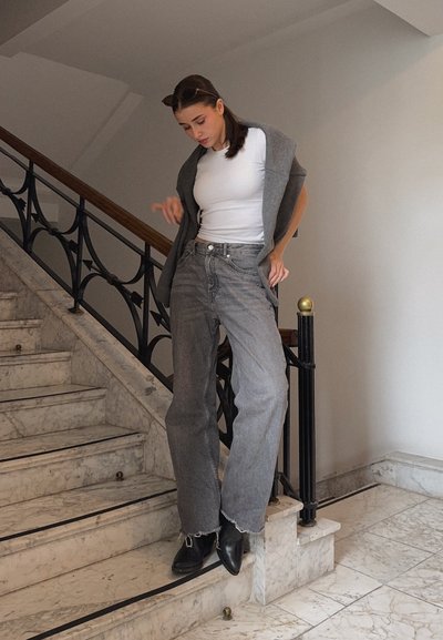 Jeans de pernera ancha grises, top ajustado blanco y un cárdigan gris sobre los hombros. Botines negros y una escalera de mármol en un ambiente luminoso.