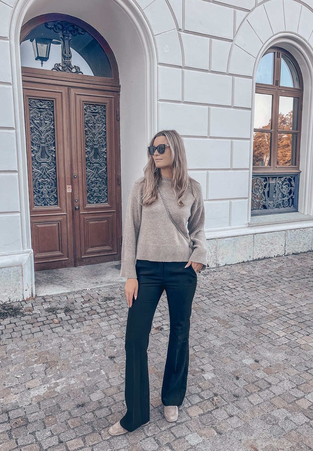 Kvinde iført solbriller, beige sweater, sorte bukser og beige sko, der står på brostensbelagt gade ved en hvid bygning med en udsmykket trædør og buet vindue.
