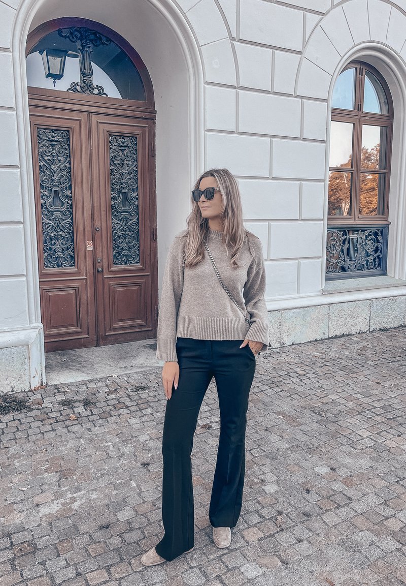 Žena koja nosi sunčane naočale, bež pulover, crne hlače i bež cipele, stoji na kockastom ulici pored bijele zgrade s ukrašenim drvenim vratima i zaobljenim prozorom.