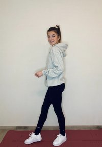 Light grey hoodie, black leggings, white trainers. Standing on a red mat, with a simple background and a ponytail hairstyle.
