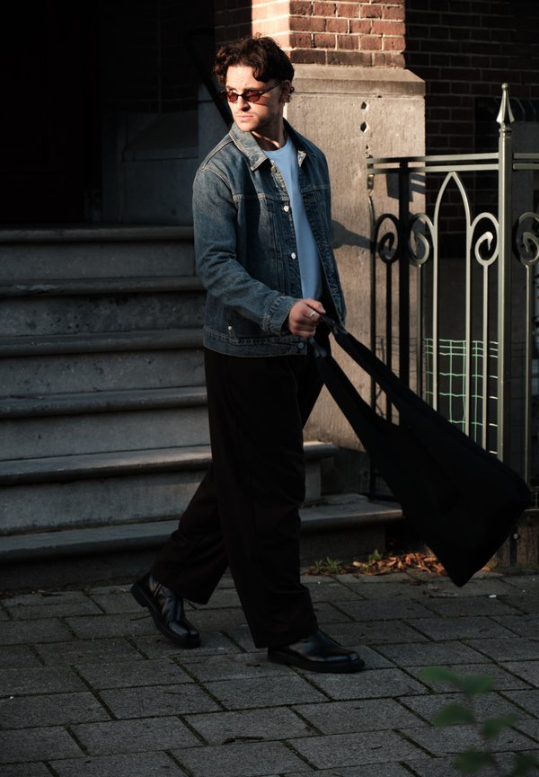 Un hombre con gafas de sol, chaqueta vaquera y pantalones negros pasa junto a unos escalones de piedra, llevando una gran bolsa negra al lado de un edificio de ladrillo.