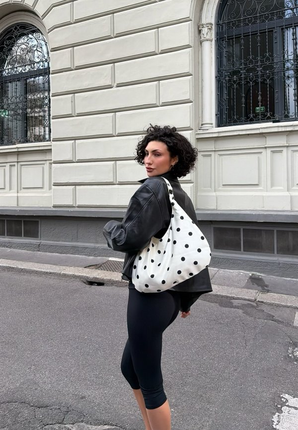 Femme aux cheveux bouclés portant une veste en cuir noire et des leggings, avec un sac à bandoulière blanc à pois, debout sur un trottoir en ville.