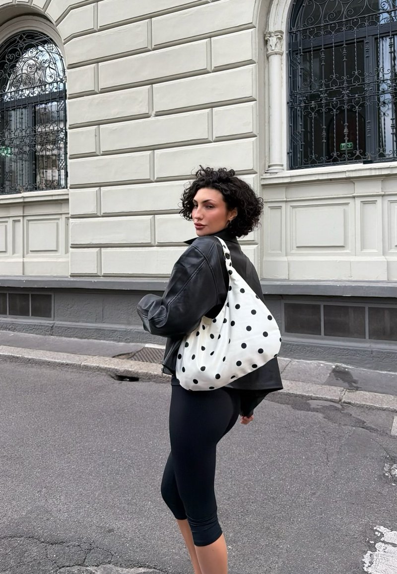 Femme aux cheveux bouclés portant une veste en cuir noire et des leggings, avec un sac à bandoulière blanc à pois, debout sur un trottoir en ville.