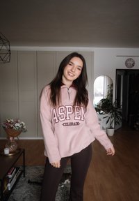 Γυναίκα με χαμόγελο, φορώντας ροζ φούτερ Aspen Colorado και σκούρο παντελόνι, στέκεται σε φωτεινό σαλόνι με φυτά, λουλούδια και μια γάτα στο πάτωμα.