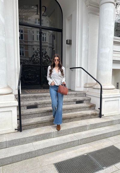 Mujer con blusa gris cruzada, jeans azules, botas marrones y gafas de sol, sosteniendo un bolso marrón, caminando por escalones de piedra junto a una puerta negra con detalles ornamentales.