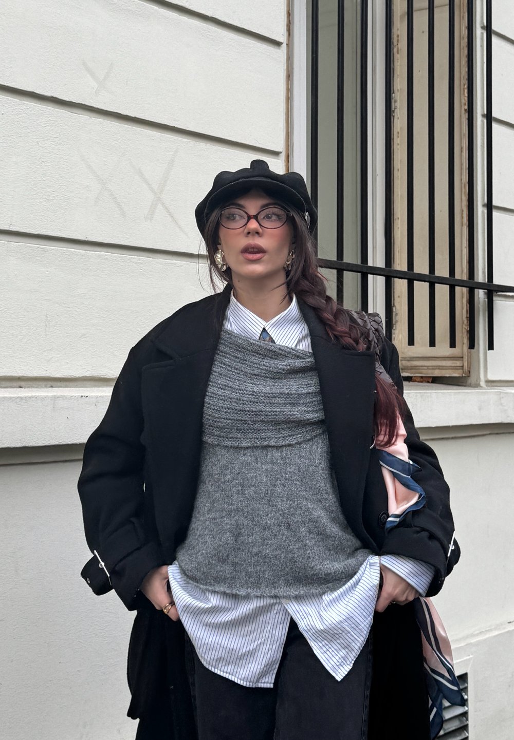 Femme avec une longue tresse portant un béret noir, des lunettes, un manteau noir, un gilet en tricot gris, une chemise rayée, debout devant un bâtiment blanc avec une fenêtre à barreaux.