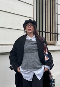 Femme avec une longue tresse portant un béret noir, des lunettes, un manteau noir, un gilet en tricot gris, une chemise rayée, debout devant un bâtiment blanc avec une fenêtre à barreaux.