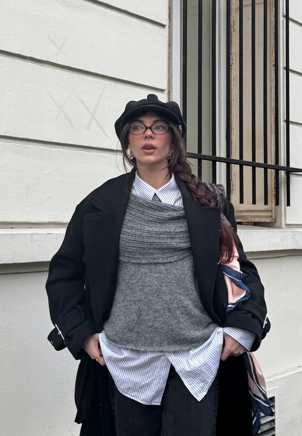 Femme avec une longue tresse portant un béret noir, des lunettes, un manteau noir, un gilet en tricot gris, une chemise rayée, debout devant un bâtiment blanc avec une fenêtre à barreaux.