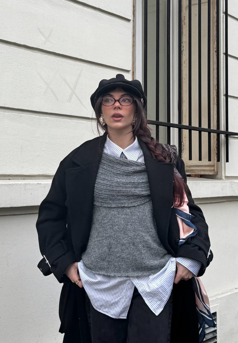 Femme avec une longue tresse portant un béret noir, des lunettes, un manteau noir, un gilet en tricot gris, une chemise rayée, debout devant un bâtiment blanc avec une fenêtre à barreaux.