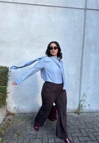 Camisa de mangas longas azul-clara com colarinho, combinada com calças largas castanhas escuras e sapatos de salto bordeaux. Os acessórios incluem óculos de sol.