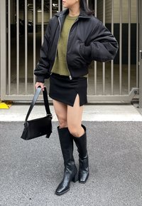 Casaco bomber preto, suéter verde-oliva, minissaia preta com fenda lateral e botas pretas de salto bloco até ao joelho. Segura uma mala preta de ombro.