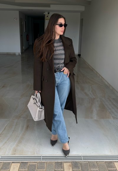 Mujer con abrigo oscuro, camisa de rayas, vaqueros azules, gafas de sol negras y tacones puntiagudos grises sosteniendo un bolso gris claro en un pasillo.