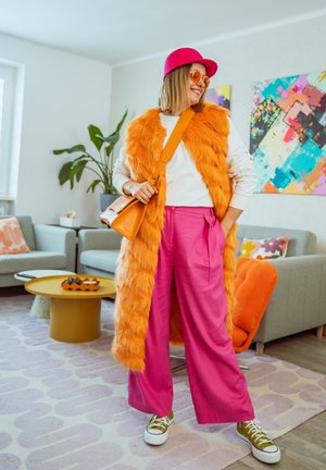 Mujer con chaleco de peluche naranja, pantalones anchos rosas, gorra rosa y gafas de sol naranjas, de pie en una sala de estar decorada con colores vivos.