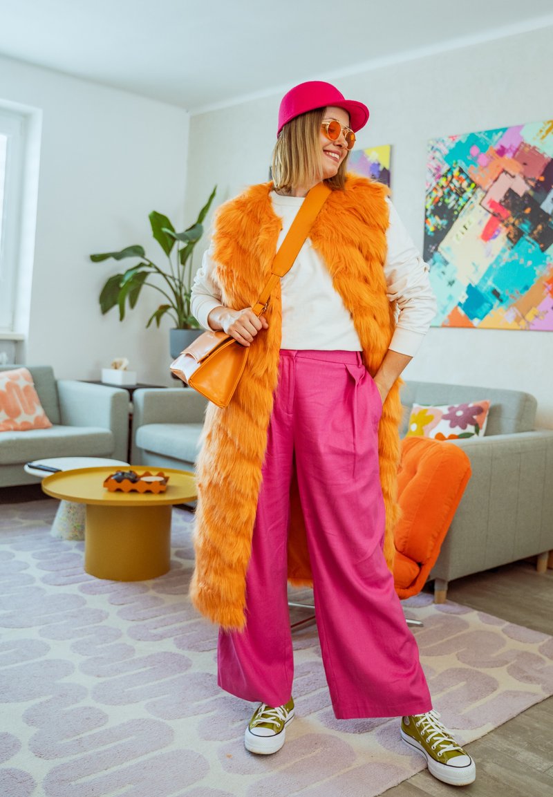 Femme portant un gilet en fausse fourrure orange, un pantalon large rose, une casquette rose et des lunettes de soleil orange, debout dans un salon décoré de manière éclatante.