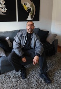 Mees prillidega ja halli jakiga istub hallil diivanil, millel on padjad, tema taga seinal on abstraktsed maalikunstiteosed.