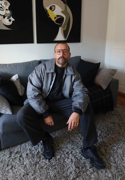 Hombre con gafas y chaqueta gris sentado en un sofá gris con cojines, pinturas abstractas en la pared detrás de él.