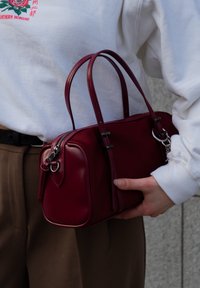 Μπορντό δερμάτινη τσάντα χειρός με διπλούς επάνω ιμάντες, ορθογώνιο σχήμα, κλείσιμο με φερμουάρ και ασημένιες λεπτομέρειες. Υφή με διακοσμητική ραφή.