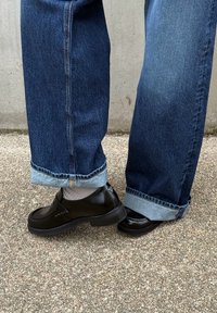 Personne portant un jean bleu foncé retroussé, des chaussettes blanches et des mocassins noirs brillants, debout sur un trottoir texturé près d'un mur en béton.
