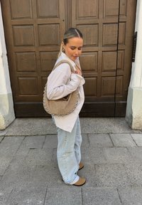 Beige Wildlederhandtasche mit Nieten und runder Form, kombiniert mit einem gestreiften oversized Hemd und hellblauen Jeans. Flache braune Schuhe auf dem Bürgersteig.